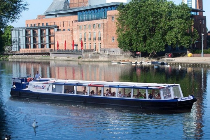 Boat on a river with buildings and trees in the background on a sunny day.