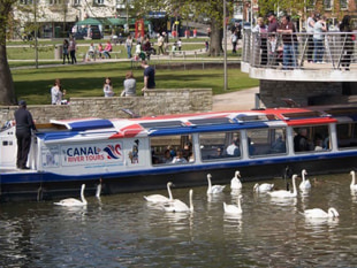 Tour boat on canal with swans, bridge, and park visitors nearby.