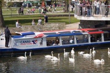 Tour boat on canal with swans, bridge, and park visitors nearby.