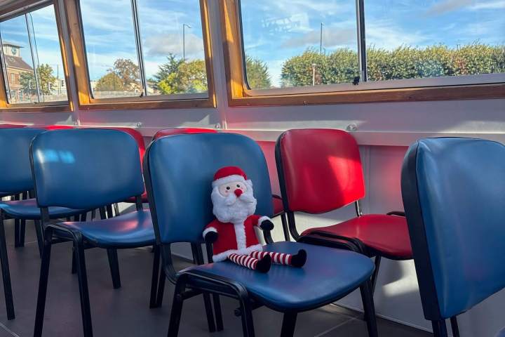 Santa doll on a blue chair in a sunlit room with red and blue chairs.